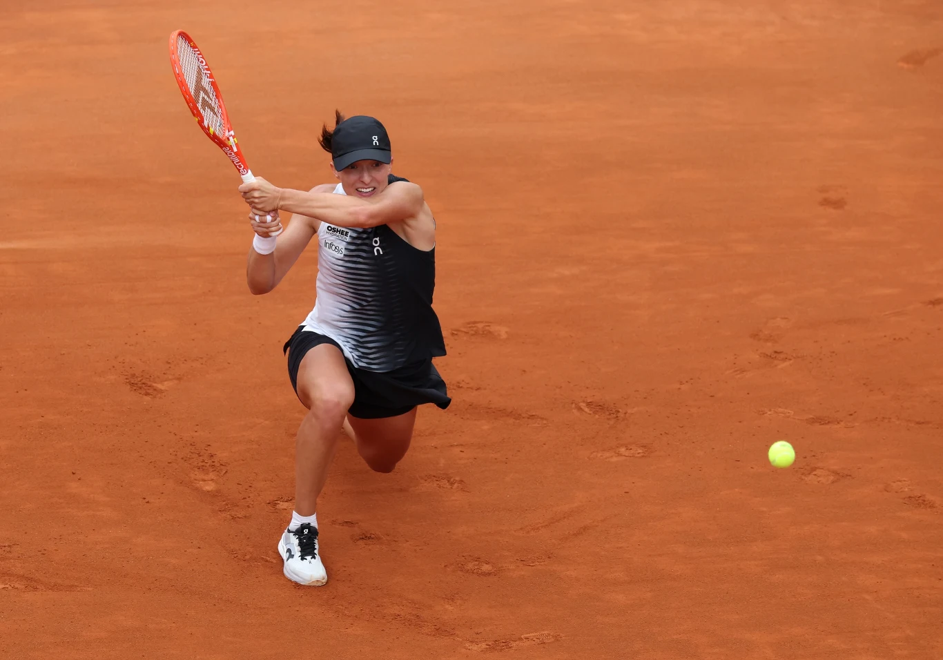 Tenisistka w sportowym stroju wykonuje dynamiczne uderzenie forhendowe na kortach ziemnych, skupiona na odbiciu nadlatującej piłki.