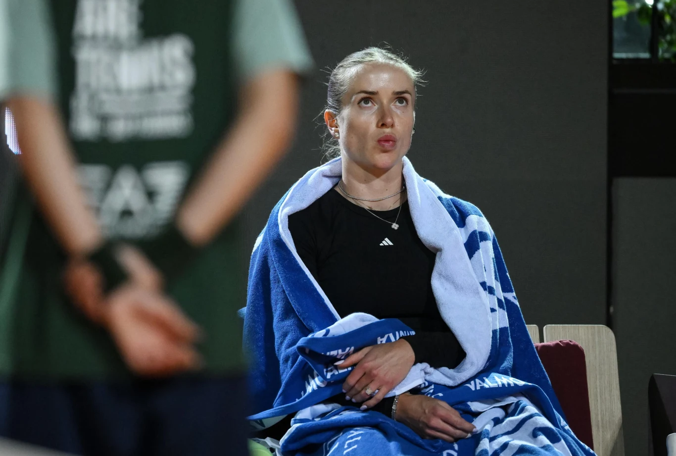 Kobieta w sportowym stroju siedząca z owiniętym wokół ramion niebieskim ręcznikiem, skupiona i zamyślona, na pierwszym planie widoczna sylwetka innej osoby stojącej tyłem.