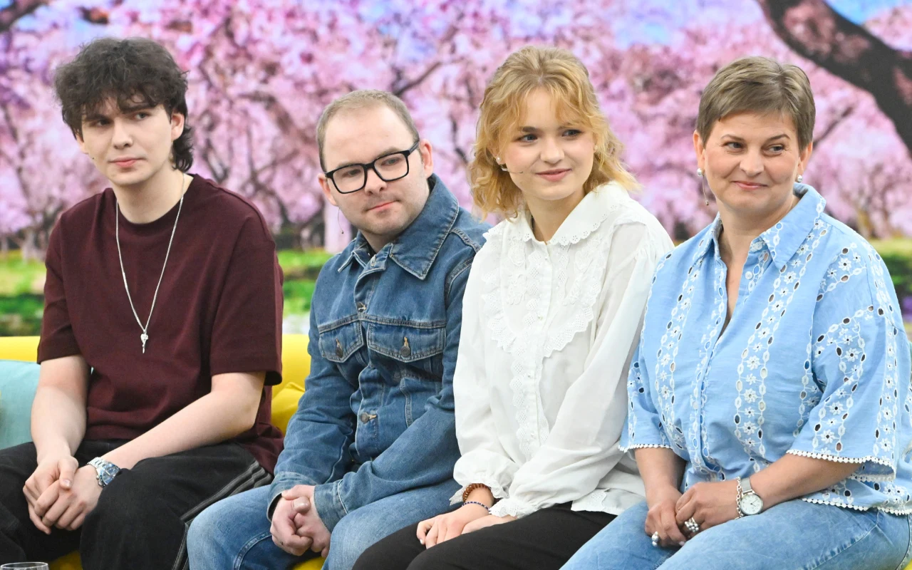 Czwórka osób siedzących na żółtej sofie, uśmiechnięci, ubrani w codzienne, wygodne stroje, na tle wiosennych drzew kwitnących na różowo.