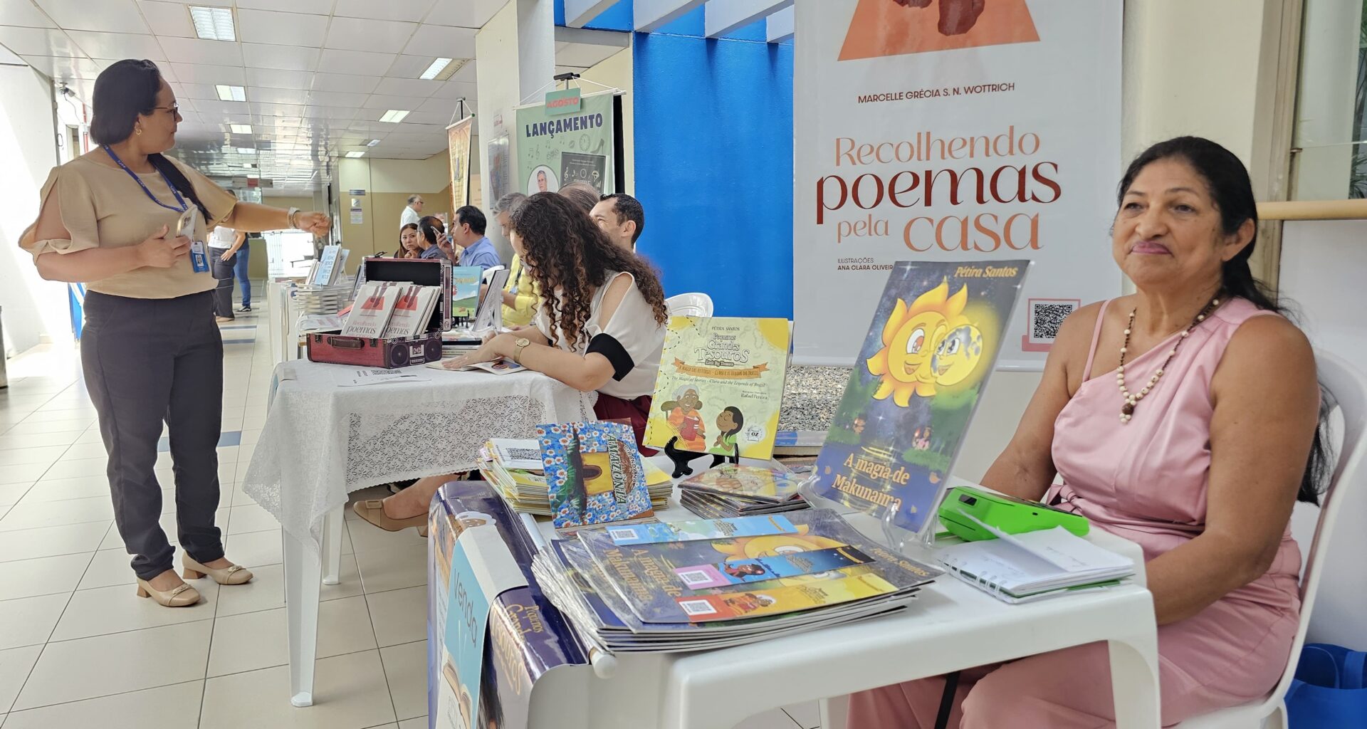 Foto colorida tirada em um corredor iluminado por luz natural e artificial, com mesas organizadas para uma feira de livros ou exposição literária, possivelmente em ambiente institucional. À direita da imagem, uma mulher de vestido rosa e colar de pérolas está sentada atrás de uma mesa com livros infantis. Ao fundo, outras mesas com autores e obras estão dispostas ao longo do corredor. Uma mulher de blusa bege e calça escura conversa com os expositores. Acima, placas de sinalização indicam que o local é próximo da 1ª Vara da Família e de sanitários. A cena transmite um ambiente de valorização da literatura local, com destaque para obras infantis e poesia.