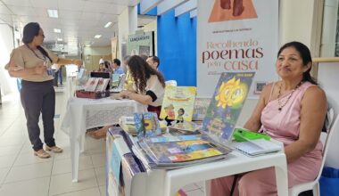 Foto colorida tirada em um corredor iluminado por luz natural e artificial, com mesas organizadas para uma feira de livros ou exposição literária, possivelmente em ambiente institucional. À direita da imagem, uma mulher de vestido rosa e colar de pérolas está sentada atrás de uma mesa com livros infantis. Ao fundo, outras mesas com autores e obras estão dispostas ao longo do corredor. Uma mulher de blusa bege e calça escura conversa com os expositores. Acima, placas de sinalização indicam que o local é próximo da 1ª Vara da Família e de sanitários. A cena transmite um ambiente de valorização da literatura local, com destaque para obras infantis e poesia.