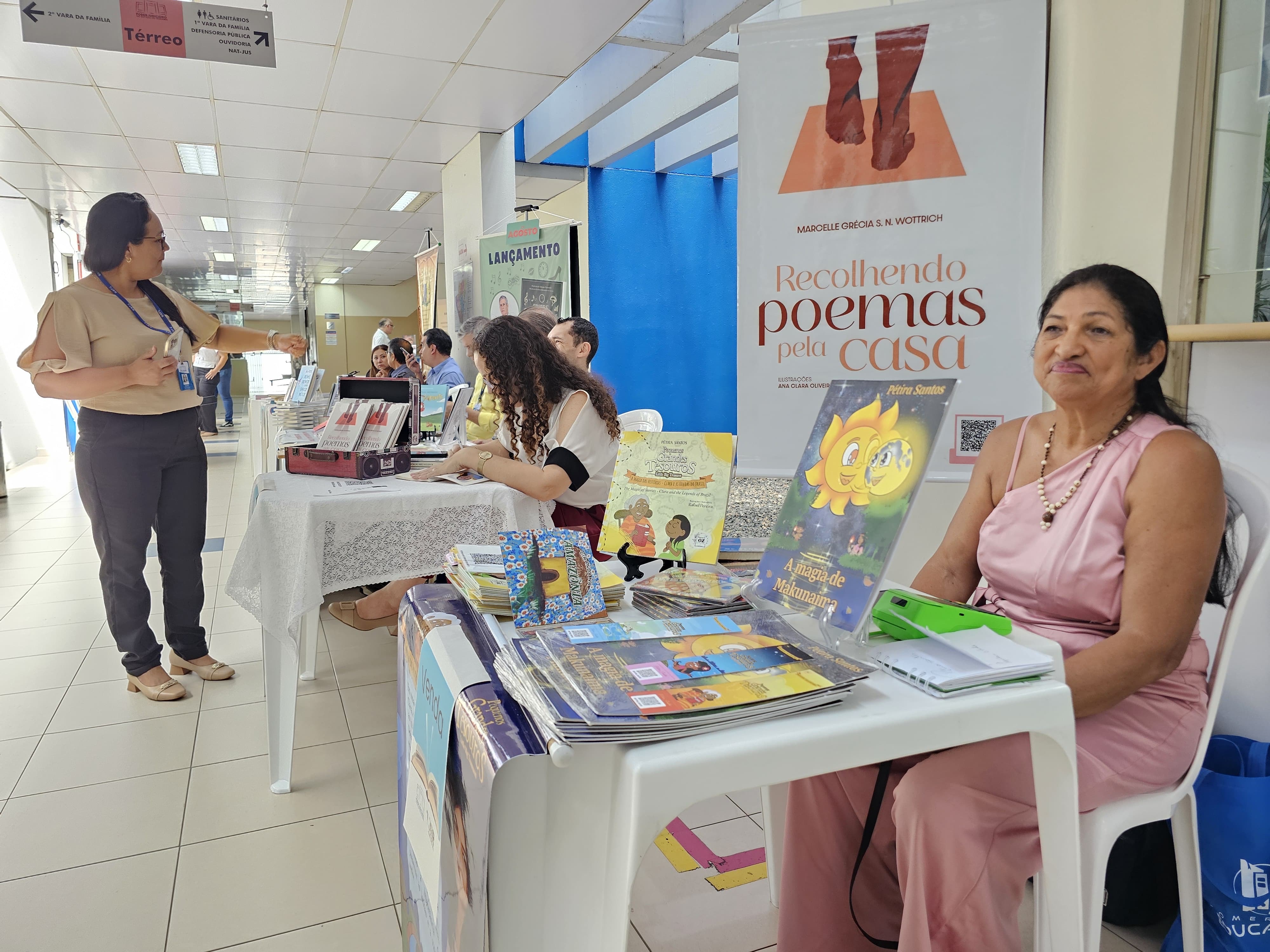  Foto colorida tirada em um corredor iluminado por luz natural e artificial, com mesas organizadas para uma feira de livros ou exposição literária, possivelmente em ambiente institucional. À direita da imagem, uma mulher de vestido rosa e colar de pérolas está sentada atrás de uma mesa com livros infantis. Ao fundo, outras mesas com autores e obras estão dispostas ao longo do corredor. Uma mulher de blusa bege e calça escura conversa com os expositores. Acima, placas de sinalização indicam que o local é próximo da 1ª Vara da Família e de sanitários. A cena transmite um ambiente de valorização da literatura local, com destaque para obras infantis e poesia.