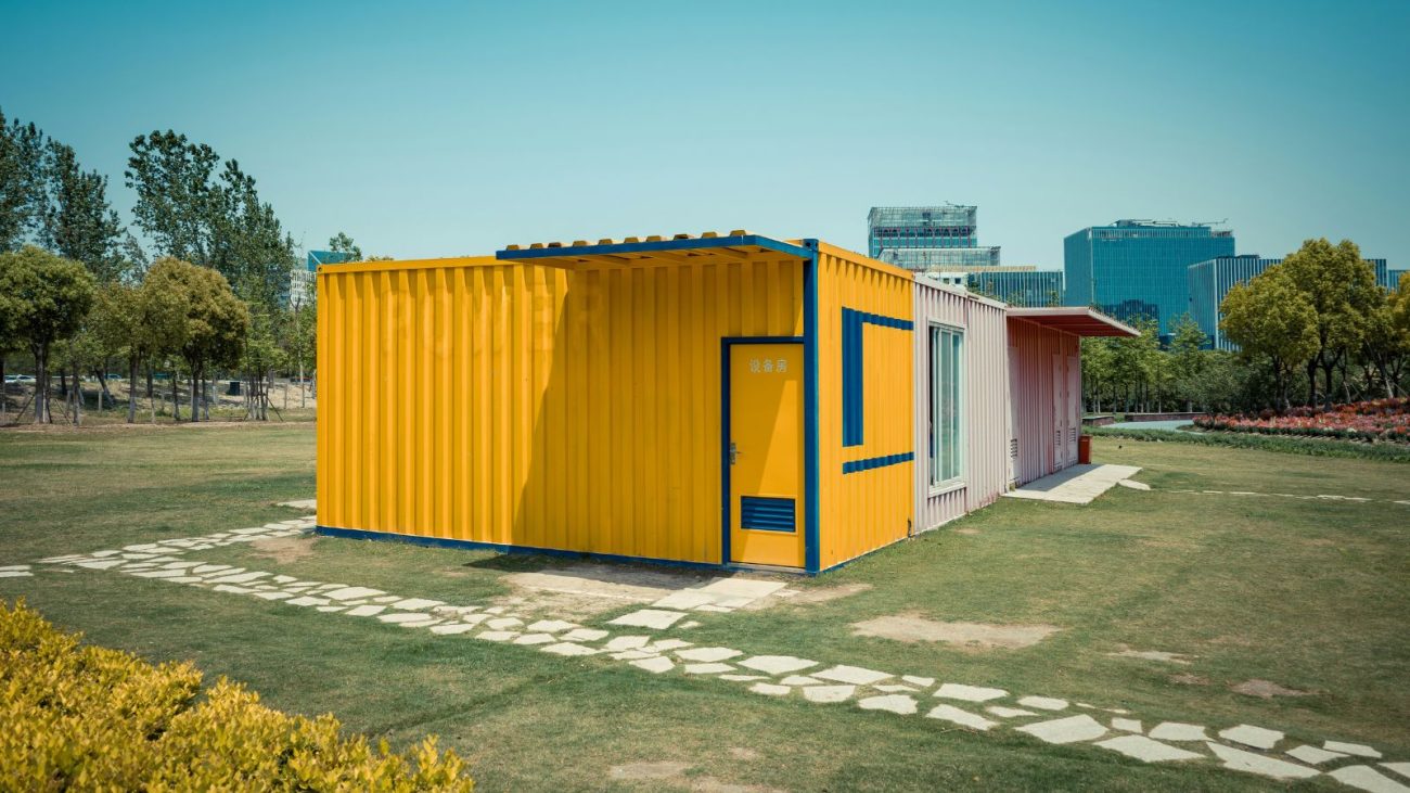 Casa moderna feita com container amarelo em área verde aberta, com caminho de pedras na entrada e edifícios ao fundo