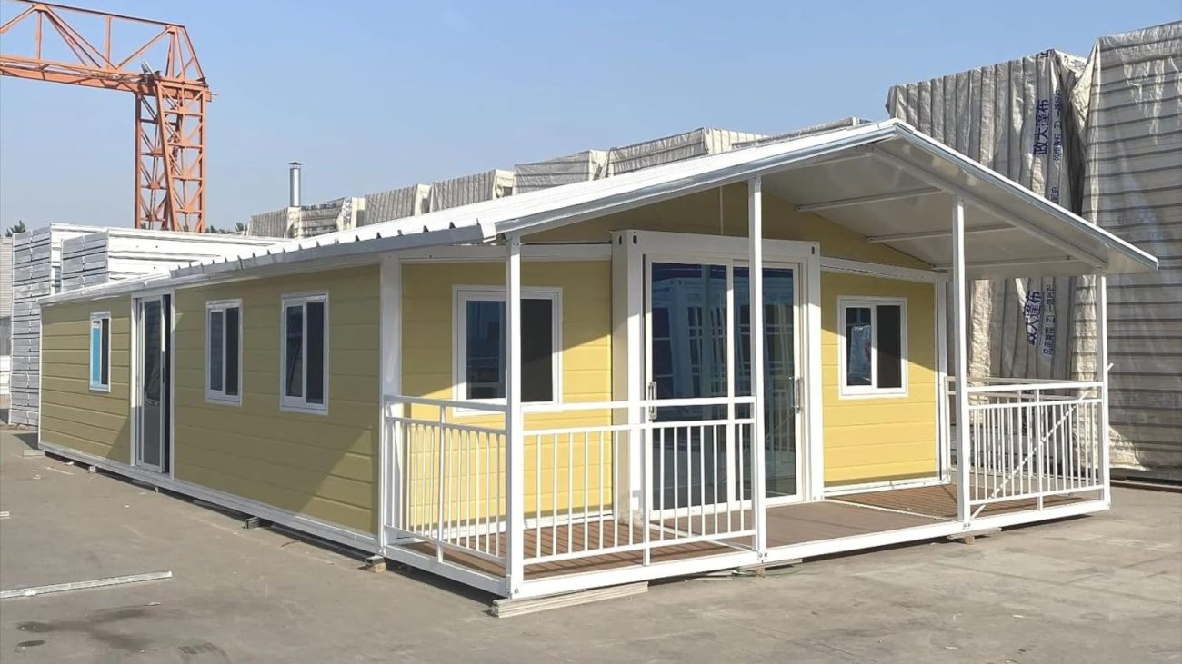 Casa da Amazon pré-fabricada de cor amarela com varanda coberta e janelas de vidro, montada em área externa e pronta para instalação em terreno residencial.