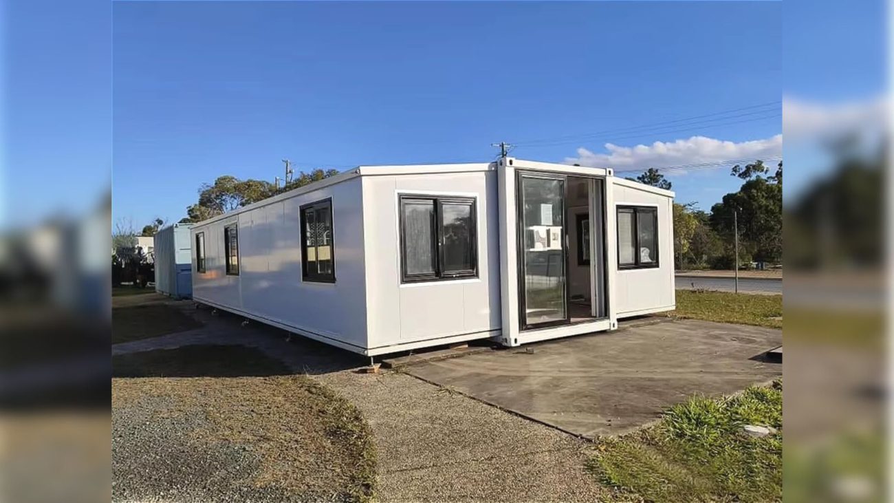  Casa da Amazon branca com estrutura de contêiner, janelas pretas e porta de vidro, instalada sobre base de concreto em ambiente aberto com gramado e céu limpo.