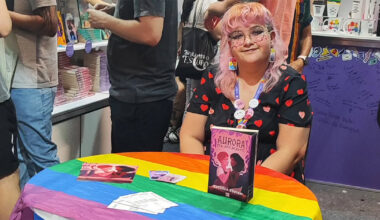 Jovem de cabelos coloridos usando óculos e um vestido estampado diante do livro Aurora Sol Assurgente