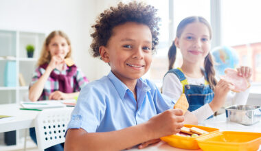 Três crianças sentadas em uma sala de aula clara e iluminada. A criança em primeiro plano, com camiseta azul, sorri enquanto segura um biscoito, com um pote de lanche amarelo à frente. Ao fundo, duas meninas, uma de macacão jeans e outra com camisa xadrez, também estão comendo.
