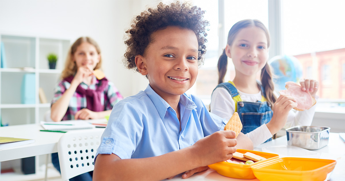 Três crianças sentadas em uma sala de aula clara e iluminada. A criança em primeiro plano, com camiseta azul, sorri enquanto segura um biscoito, com um pote de lanche amarelo à frente. Ao fundo, duas meninas, uma de macacão jeans e outra com camisa xadrez, também estão comendo.
