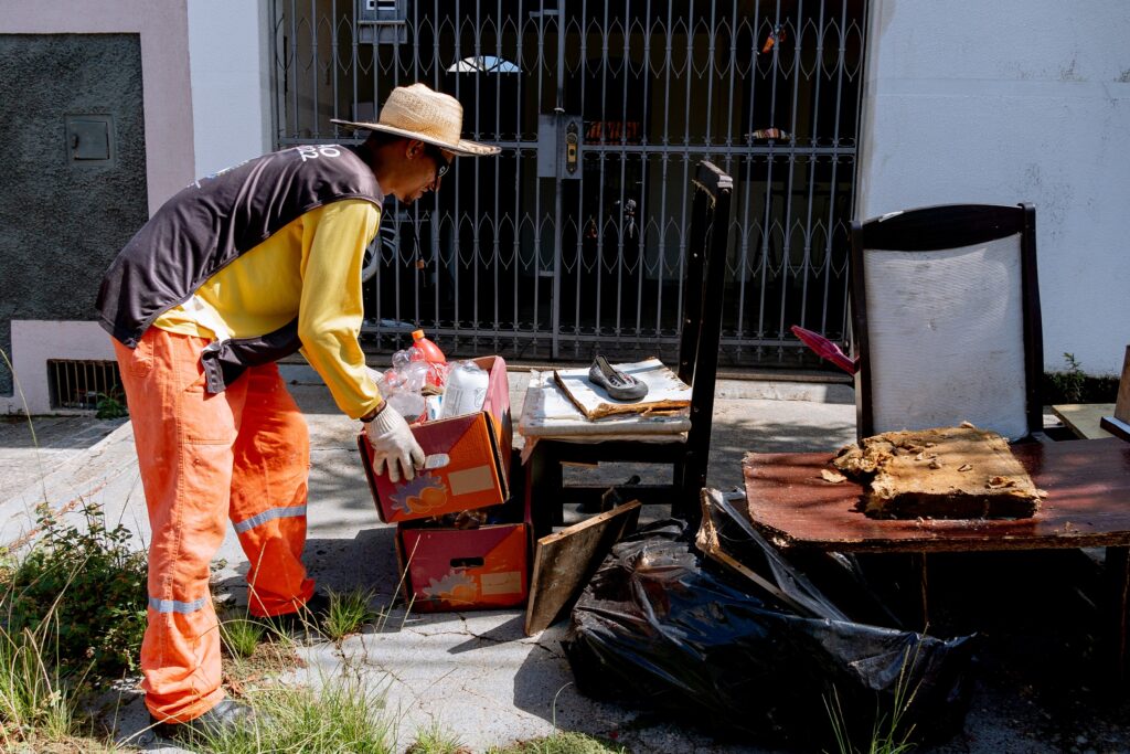 Um trabalhador com um chapéu de palha, óculos escuros, camisa amarela, colete preto e laranja, e luvas brancas, está carregando duas caixas de papelão cheias de garrafas plásticas e outros itens. As caixas estão empilhadas uma sobre a outra. Ele está em frente a um portão de metal gradeado, com uma casa ao fundo. Há também uma cadeira de madeira com um sapato em cima, uma mesa com um pedaço de espuma e um saco de lixo preto no chão.