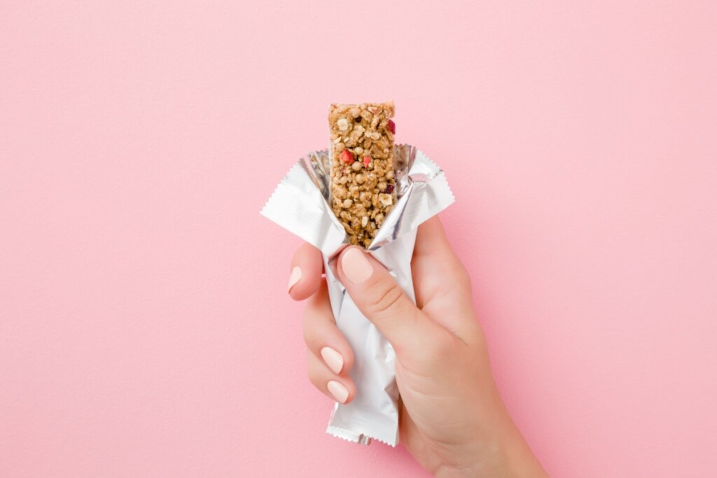 Uma mÃ£o segurando uma barrinha de cereal com a embalagem aberta em fundo rosa