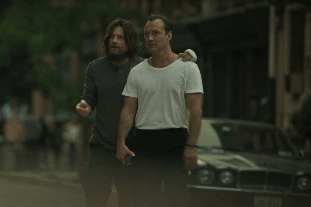 Dois homens juntos em uma rua, um deles com camiseta branca e o outro com barba e cabelo comprido usando camisa escura