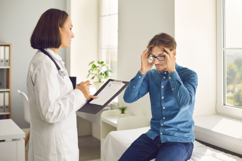 MÃ©dica em consultÃ³rio avaliando paciente, que estÃ¡ com as mÃ£os na cabeÃ§a, sugerindo enxaqueca
