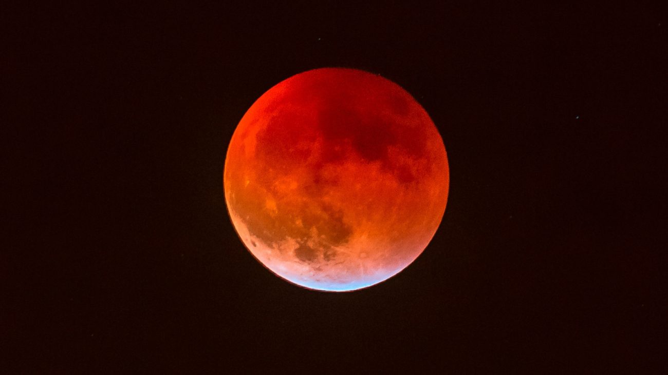 Lua vermelho sangue durante um eclipse lunar, com tonalidades intensas de vermelho e laranja, no céu escuro com algumas estrelas visíveis.