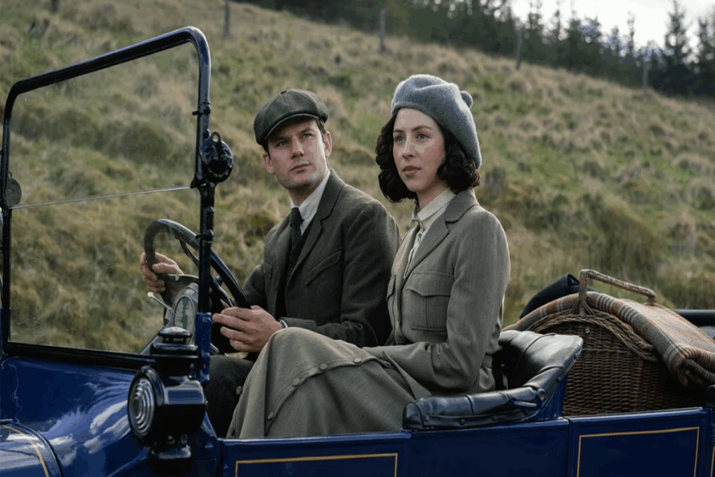 Um casal vestindo roupas de inverno de época sentados dentro de um carro com uma montanha ao fundo