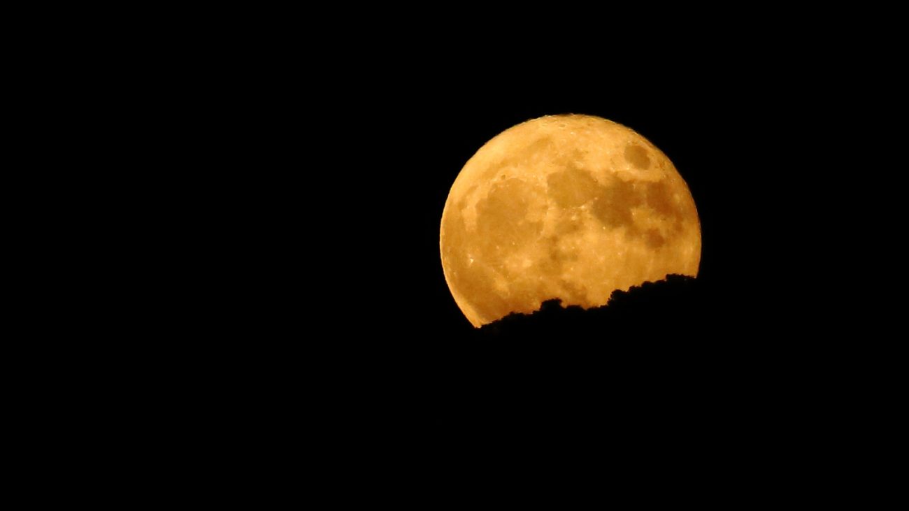 Imagem da lua cheia com tom dourado, visível no céu noturno, parcialmente coberta por uma silhueta de montanha. Perfeita para observação astronômica e céu noturno.