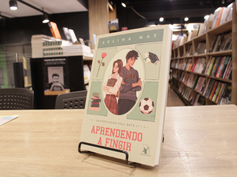 Livro "Aprendendo a fingir", da autora Selina Mae, em uma mesa com livros em estante ao fundo.