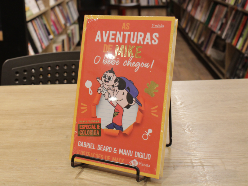 Livro "Aventuras de Mike 2: O bebê chegou!" dos autores Gabriel Dearo e Manu Digilio em uma mesa com livros em estante ao fundo.