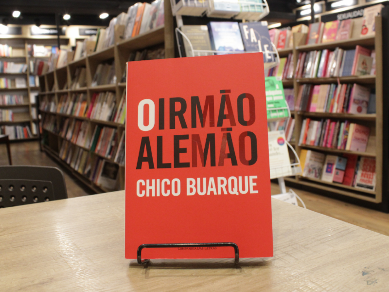 Livro "O Irmão Alemão", do autor Chico Buarque, em uma mesa com livros em estante ao fundo.