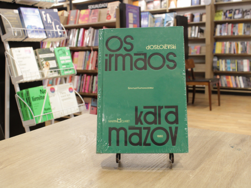 Livro "Irmãos Karamazov", do autor Fiódor Dostoiévski, em uma mesa com livros em estante ao fundo.