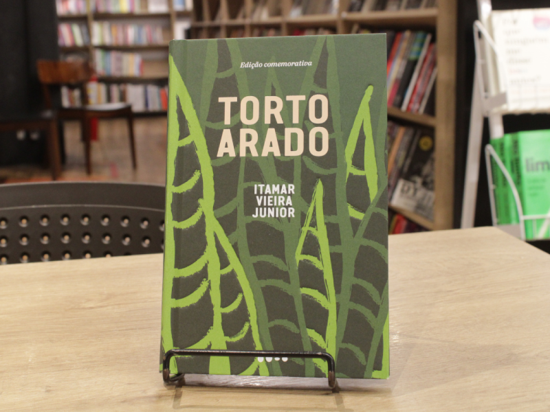 Livro "Torto Arado", do autor Itamar Vieira Junior, em uma mesa com livros em estante ao fundo.