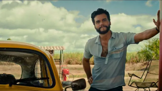Cena de O Agente Secreto, com o ator Wagner Moura de pé, vestindo uma camisa azul, ao lado de um fusca amarelo.
