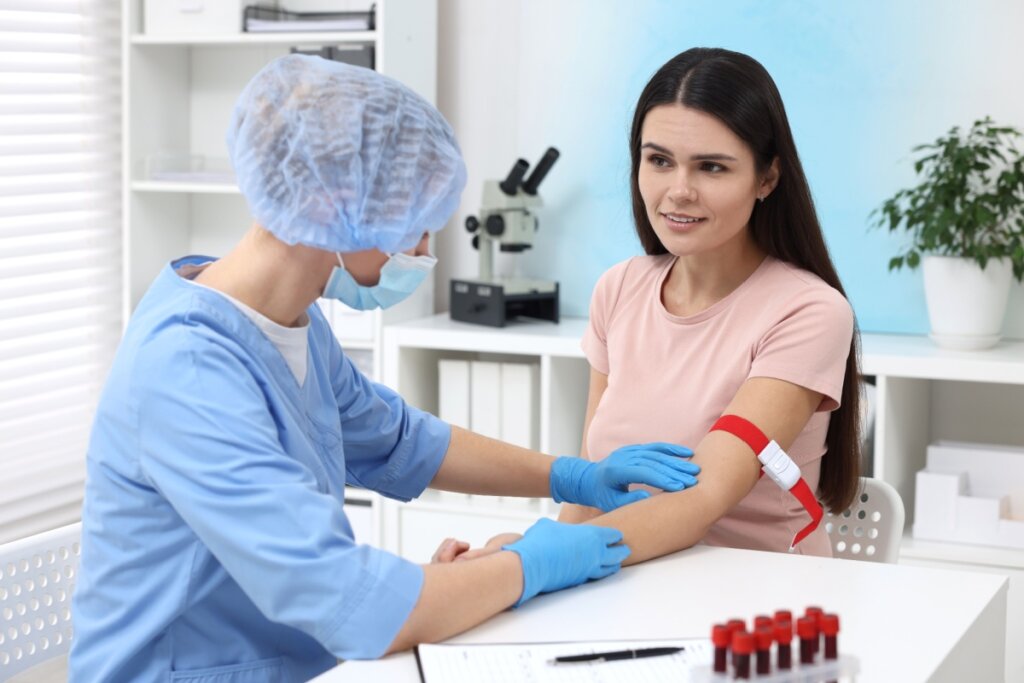 Profissional de saúde com máscara e touca preparando braço de paciente para coleta de sangue