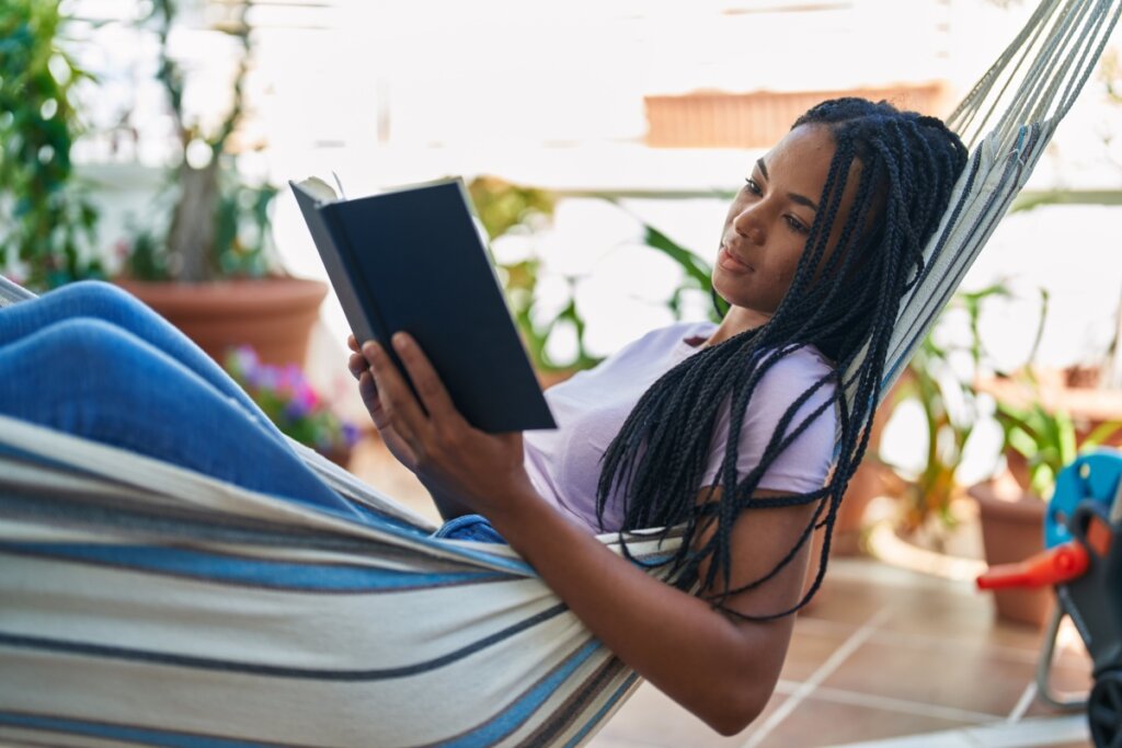 Jovem com o cabelo longo, tranÃ§ado, usando uma camiseta roxa deitada em rede listrada no quintal com um livro na mÃ£o