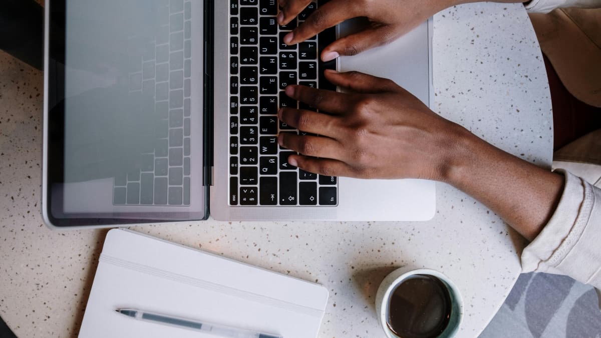 Uma pessoa a tirar notas: veem-se umas mãos em posição de digitação sobre um teclado de um computador portátil. Ao lado, um café e uma bloco de notas físico.