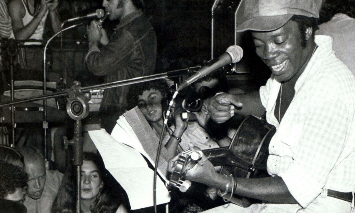 Milton Nascimento está no palco e toca violão, sorrindo e sentado em banco, durante show realizado em 1975 em Belo Horizonte