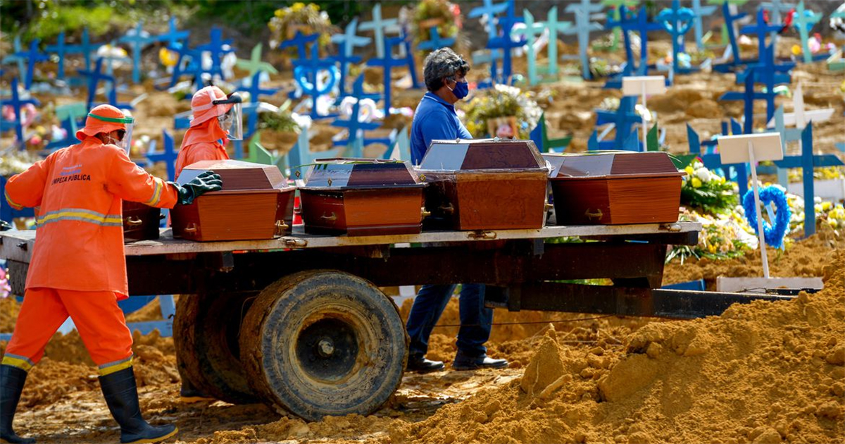 Imagem de uma carroceria de caminhão onde estão expostos vários caixões sob os cuidados de vários homens de uniforme laranja ou azul