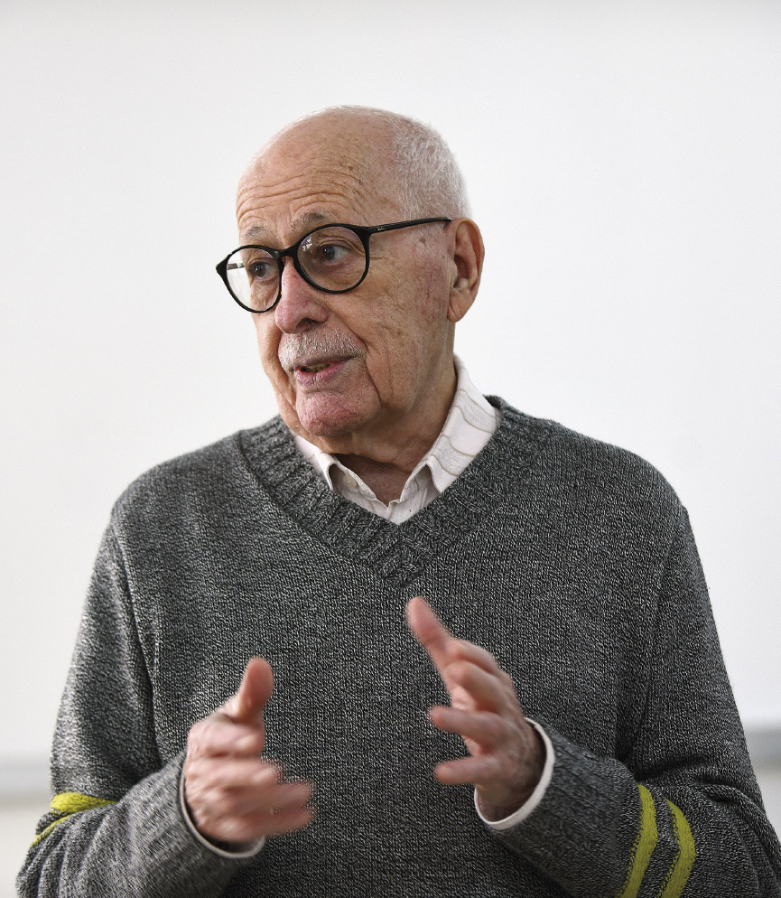 Esta imagem é um retrato em plano médio de um homem idoso, branco, com a cabeça calva e um bigode branco bem aparado. Ele usa óculos de armação escura e tem uma expressão facial engajada, com a boca ligeiramente aberta, como se estivesse no meio de uma conversa ou palestra.

Ele está gesticulando com ambas as mãos à sua frente para enfatizar um ponto. Veste um suéter de malha cinza com gola em V, por baixo do qual se vê uma camisa social. Um detalhe notável são as duas listras amarelas vibrantes nos antebraços do suéter. O fundo é liso e de cor clara, o que mantém todo o foco na figura do homem.