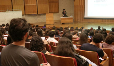 Auditório lotado de estudantes e uma pessoa no palco falando e ao fundo uma apresentação numa tela