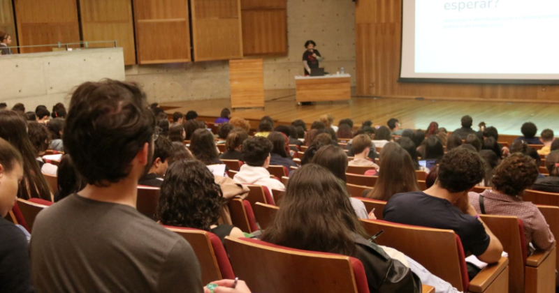 Auditório lotado de estudantes e uma pessoa no palco falando e ao fundo uma apresentação numa tela