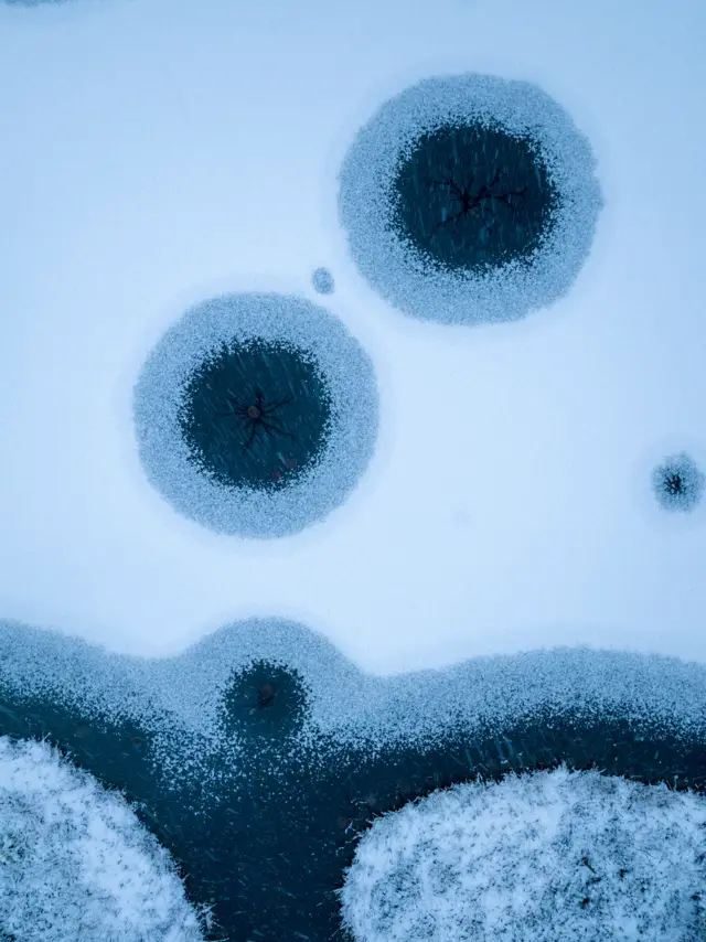 Superfície de lago congelado mostra três círculos escuros rodeados por gelo rachado. Ao fundo, linhas curvas maiores de neve e gelo rodeiam os círculos.