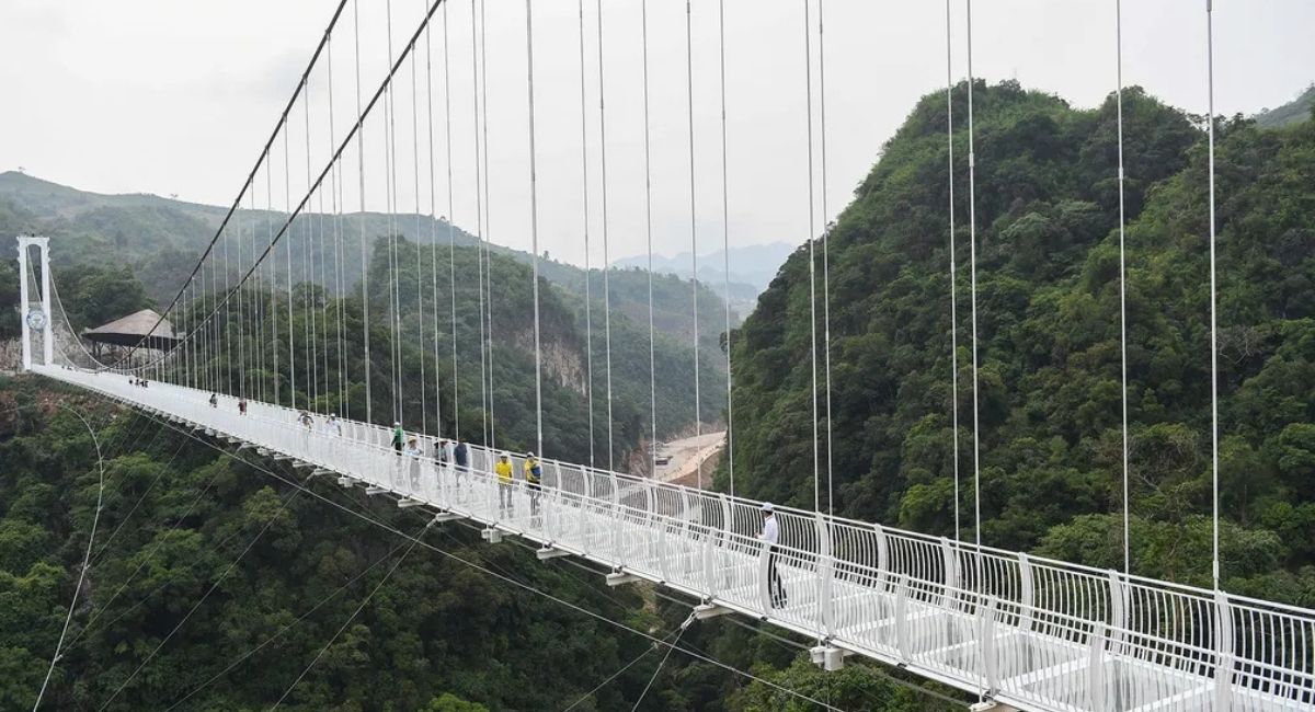 Turistas caminham sobre a maior ponte de vidro do mundo, suspensa entre montanhas cobertas por vegetação densa, com vista panorâmica para um vale profundo.