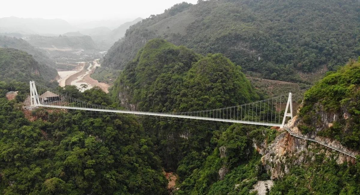 Vista aérea da maior ponte de vidro do mundo, ligando dois grandes penhascos cobertos por floresta, com um rio serpenteando no fundo do vale.