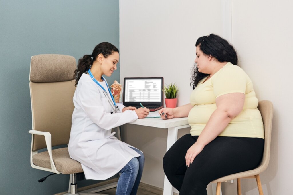 imagem de médica de jaleco branco e paciente obesa de camiseta amarela sentadas em cadeiras em consultório