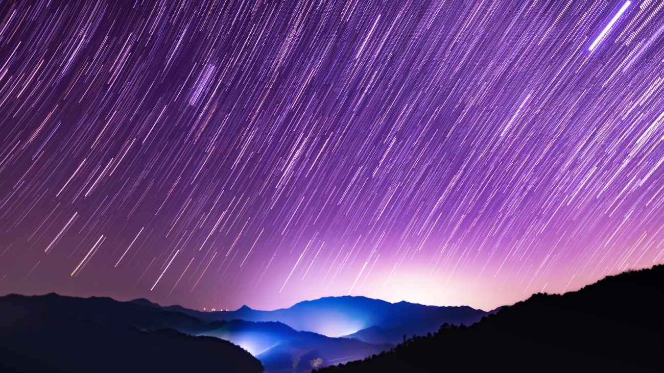 Céu noturno estrelado com trilhas de estrelas visíveis sobre montanhas com cores violetas e azuis, exibindo um espetáculo celestial impressionante.