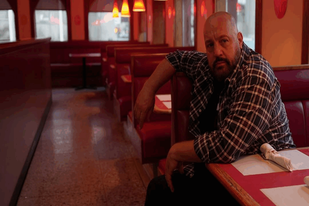 Homem careca e com barba sentado em mesa de lanchonete usando camisa xadrez