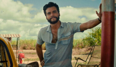 Homem de camisa azul clara encostado em bomba de combustível vermelha em posto ao ar livre. Carro amarelo estacionado à esquerda, céu azul com nuvens ao fundo.