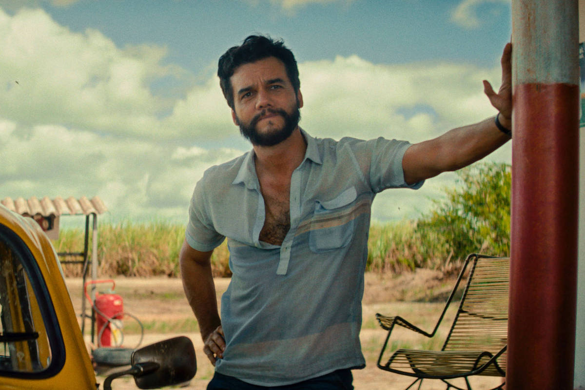 Homem de camisa azul clara encostado em bomba de combustível vermelha em posto ao ar livre. Carro amarelo estacionado à esquerda, céu azul com nuvens ao fundo.