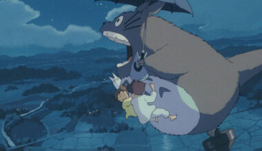 Totoro, criatura grande e cinza, voa segurando um guarda-chuva aberto à noite. Três crianças estão penduradas em seu corpo enquanto sobrevoam uma paisagem rural iluminada por luzes distantes.