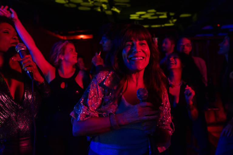 Grupo de pessoas em ambiente escuro com iluminação colorida, algumas segurando microfones e outras dançando. Mulher ao centro sorri segurando microfone, vestindo blusa brilhante. Fundo com mais pessoas em movimento e luzes decorativas no teto.
