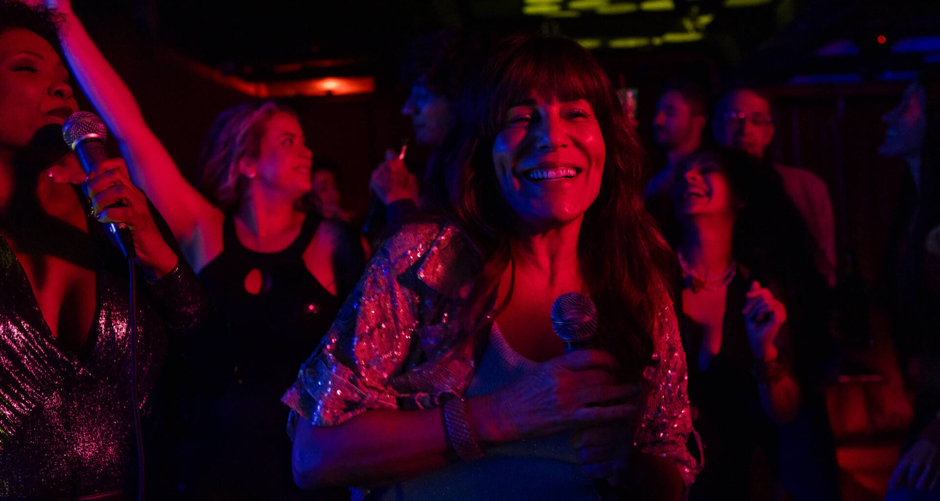 Grupo de pessoas em ambiente escuro com iluminação colorida, algumas segurando microfones e outras dançando. Mulher ao centro sorri segurando microfone, vestindo blusa brilhante. Fundo com mais pessoas em movimento e luzes decorativas no teto.