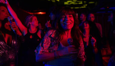Grupo de pessoas em ambiente escuro com iluminação colorida, algumas segurando microfones e outras dançando. Mulher ao centro sorri segurando microfone, vestindo blusa brilhante. Fundo com mais pessoas em movimento e luzes decorativas no teto.
