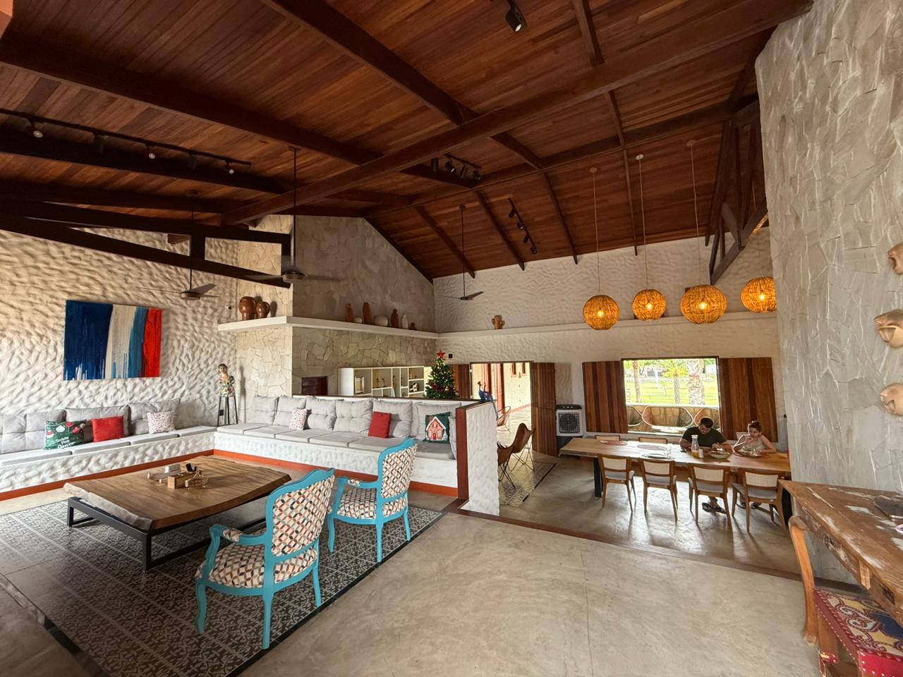 Na imagem, uma sala de estar ampla e rústica com pé-direito alto e teto inclinado de madeira escura com vigas expostas. As paredes são revestidas com pedras naturais em tons de bege claro, criando uma textura orgânica. No centro da sala, há um tapete com estampa geométrica em preto e branco sob uma mesa de centro robusta de madeira maciça. Ao fundo, um longo sofá embutido com base de alvenaria branca e estofamento cinza claro percorre a lateral da sala. Sobre a parede de pedra à direita, uma composição artística com várias pequenas cerâmicas arredondadas está fixada, acima de um aparador de madeira. A decoração inclui elementos de arte popular e regional, como esculturas de cabeças de barro e uma estátua colorida. No fundo do ambiente, é possível ver uma pequena árvore de Natal decorada e janelas de madeira que se abrem para uma área externa verde. A iluminação é composta por trilhos de spots pretos no teto e luminárias pendentes em formato de esferas de palha sobre uma mesa de jantar ao fundo.