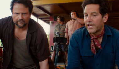 Dois homens estão agachados em primeiro plano, um com camisa preta aberta e camiseta cinza, outro com camisa azul e lenço vermelho no pescoço. Ao fundo, dois homens operam equipamentos de filmagem em área externa com estrutura de madeira.