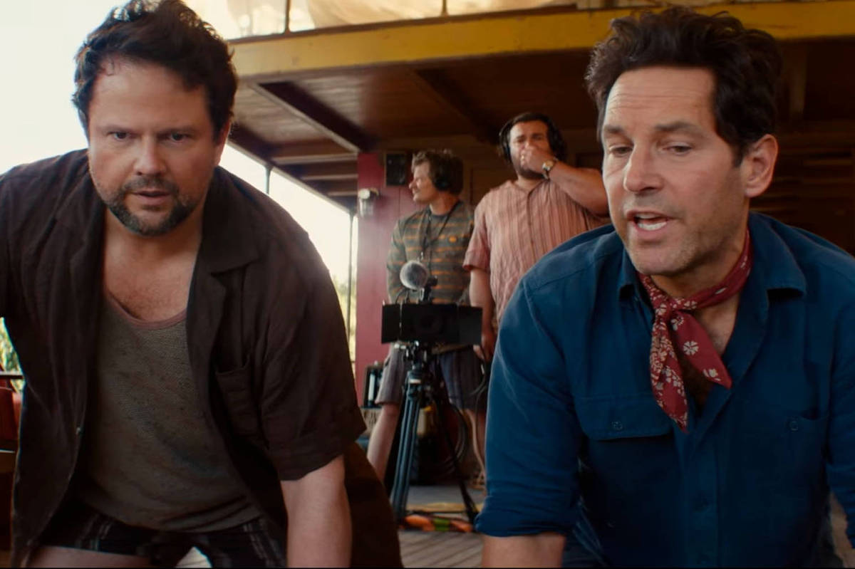 Dois homens estão agachados em primeiro plano, um com camisa preta aberta e camiseta cinza, outro com camisa azul e lenço vermelho no pescoço. Ao fundo, dois homens operam equipamentos de filmagem em área externa com estrutura de madeira.