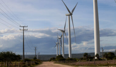 A foto mostra uma estrada de terra cercada por vegetação baixa, com vários aerogeradores (turbinas eólicas) alinhados ao lado direito e postes de energia com fios ao lado esquerdo, sob um céu parcialmente nublado. É uma paisagem rural com infraestrutura de geração e transmissão de energia.