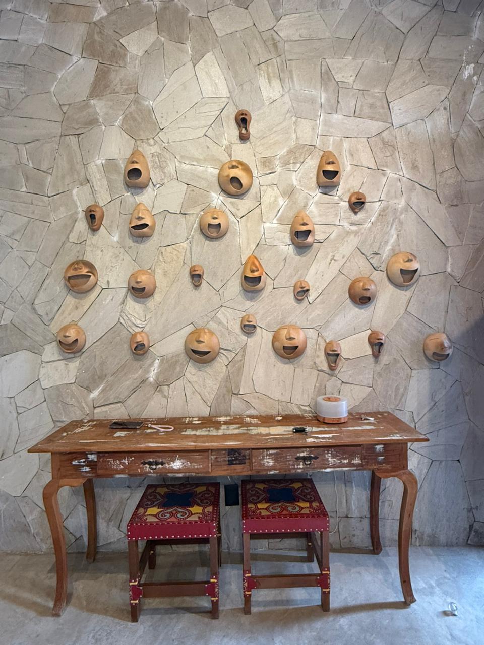 Na imagem, close-up de uma parede de destaque revestida com pedras irregulares de tom bege claro. Pendurada na parede, há uma coleção de máscaras ou cabeças de cerâmica em tons de terracota, cada uma com expressões faciais variadas, como sorrisos e bocas abertas. Abaixo da composição, encontra-se um aparador de madeira rústica com acabamento desgastado. Sob o aparador, estão guardados dois bancos (pufes) quadrados com estofamento vermelho e padrões coloridos de inspiração folclórica.